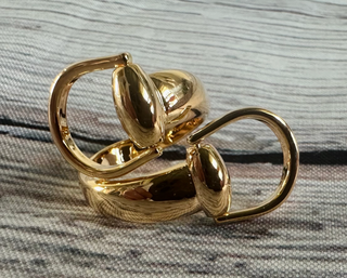 A high-polish gold-tone ring snaffle scarf slide with an interlocking snaffle bit design featuring smooth, rounded loops, photographed on a textured gray and blush fabric surface.