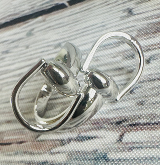 A high-polish gold-tone ring snaffle scarf slide with an interlocking snaffle bit design featuring smooth, rounded loops, photographed on a textured gray and blush fabric surface.